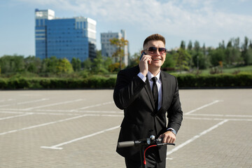 Man in a black business suit stands next to an electric scooter and talks on the phone