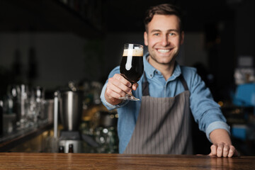 Cheers. Smiling barman holds out glass of dark beer with foam, for client