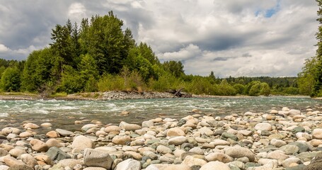 A boulder strewn, fast flowing river, beside a forest, on a cloudy day.