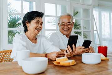 Happy elderly Asian man and woman smiling and browsing social media on smartphones while having breakfast together. Senior couple using mobile phone at home.