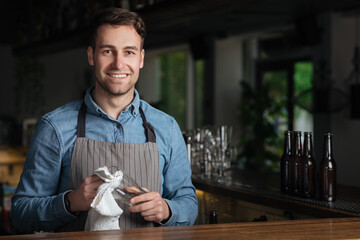 Opening pub. Smiling handsome barman in apron wipes glass