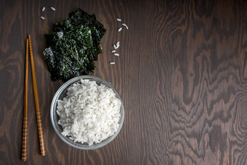Bowl of white rice, chopsticks and nori sheets are laying on dark wooden table at kitchen