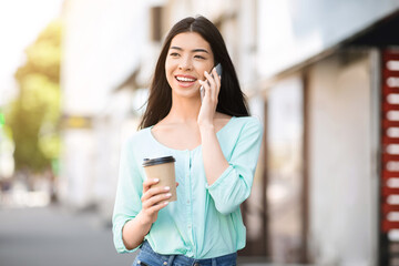 Modern Communication. Asian girl walking outdoors with coffee and talking on cellphone