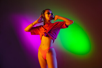 Seductive young girl' portrait on bicolored studio background in neon. Fit woman in bodysuit listen to music in headphones. Facial expression, summer, weekend, beauty, resort concept. Vacations.