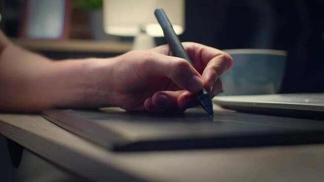 Close-up shot of young handsome man draws with a pen on a tablet at home at night at a table in the living room