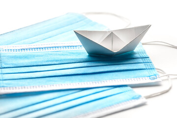 Sailing during pandemic crisis. Surgical mask with white paper ship on white background.