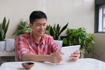  one young Asian man using tablet computer at home. Green potted plants as background