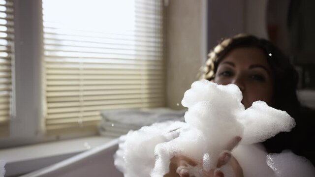 Attractive woman playing with foam in bathroom. Video. Beautiful woman blows up foam in bathroom on background of window. Sexy and fun game of women with foam