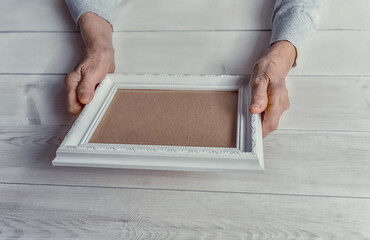 Fototapeta na wymiar Senior wrinkled hands holding white photo frame. Elderly woman looking at the picture, light wodden background. Empty template, mock-up. Happy memories, sadness, nostalgia concept.