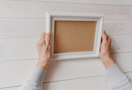 Senior wrinkled hands holding white photo frame. Elderly woman looking at the picture, light wodden background. Empty template, mock-up. Happy memories, sadness, nostalgia concept.