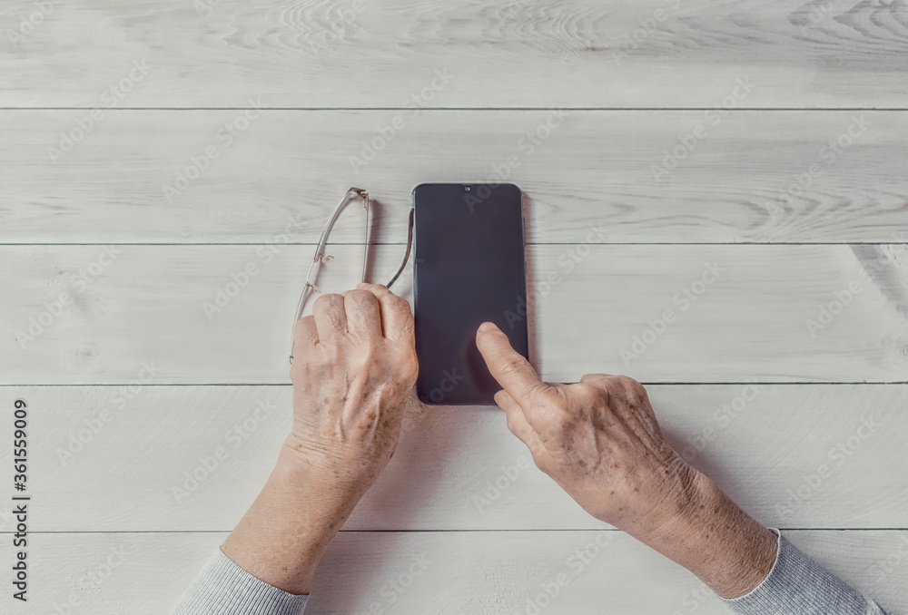 Wall mural Old wrinkled hands holding mobile smart phone. Elderly woman using digital phone showing something on display, light table. Template, copy space mockup. Teaching, technologies for seniors concept.