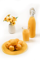 Composition on a white background with apricots in a plate, a vase with yellow flowers, a bottle and a glass of apricot juice.