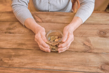 Old wrinkled hands holding jar with coins, wooden background. Saving money for future plan and retirement fund, pension, poorness, need concept.