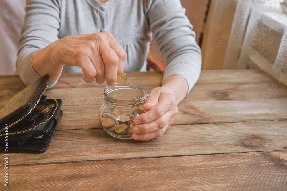 Wall mural Old wrinkled hand holding jar with coins, empty wallet, wooden background. Elderly woman throws a coin into a jar, counting. Saving money for future, retirement fund, pension, poorness, need concept.