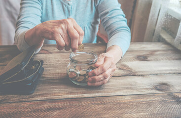 Old wrinkled hand holding jar with coins, empty wallet, wooden background. Elderly woman throws a coin into a jar, counting. Saving money for future, retirement fund, pension, poorness, need concept.