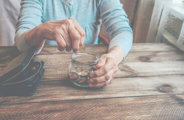 Old wrinkled hand holding jar with coins, empty wallet, wooden background. Elderly woman throws a...