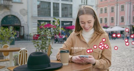 The girl is sitting at a table in a cafe and looking through their social networks. She is very happy with the number of likes. On the table a cup of coffee and a hat. The Central square of the city
