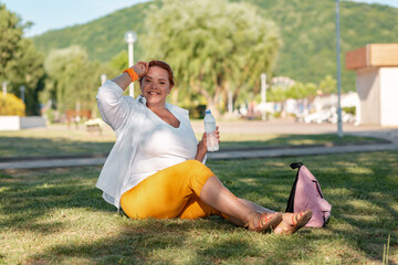 Fototapeta premium A smiling overweight woman sits on the grass in a Park and holds a water bottle. Thirst and rest after a walk