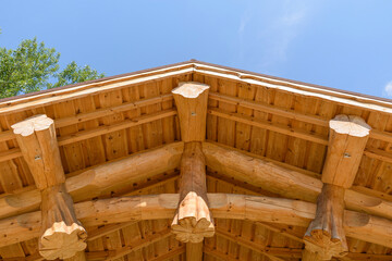 Wooden roof made of large logs and boards. Green foliage of a tree. Background is blue sky. Concept of a canopy protecting from adverse weather, a pagan temple.