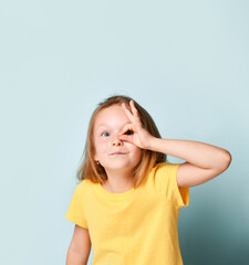 Blonde little girl in yellow t-shirt. Showing ok sign and looking through it while posing against turquoise studio background. Close up