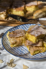 Four pieces of homemade traditional Greek pastry -- bougatsa made with phyllo dough and semolina custard cream on white and blue plate with rest of pastry, vintage spoons and lavender on white table.