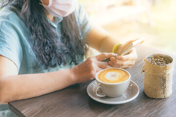 Hot cappuccino coffee for quarantined woman to prevent the spread of the Coronavirus(Covid 19). Woman wearing face mask relaxing with coffee and mobile phones. New normal. Selective focus on coffee.