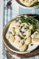 Homemade potato gnocchi with creamy gorgonzola sauce decorated with capers and arugula in white plate on wooden board.
