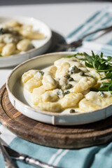 Homemade potato gnocchi with creamy gorgonzola sauce decorated with capers and arugula in white plate on wooden board.
