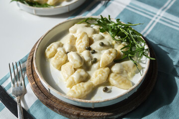 Homemade potato gnocchi with creamy gorgonzola sauce decorated with capers and arugula in white plate on wooden board. - obrazy, fototapety, plakaty