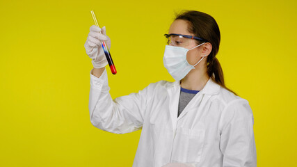 Doctor in a medical mask, goggles and latex gloves looks at the reaction in test tube. Young girl in a white coat on a yellow background holds hand a flask with red-black liquid