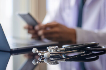 Male doctor in white lab coat, hand holding and using mobile smart phone with laptop computer and medical stethoscope on the desk. Medical online networking, telehealth, telemedicine, ehr, emr concept