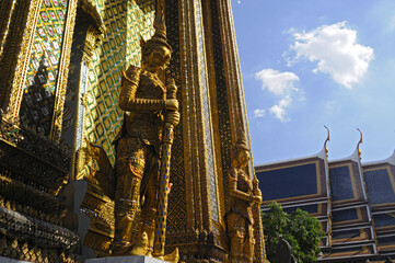 Obraz premium Vue sur deux statue dorée de guerriers protégeant l'entrée d'un temple au palais impérial de Bangkok. Mosaïque en miroir étincelant au soleil sur fond de ciel bleu.