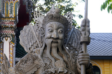 Fototapeta na wymiar Gros Plan sur le visage d'une statue de pierre représentant un guerrier du Siam, à longue barbe et longue moustache, tenant une hallebarde à l'entrée d'un temple Bouddhiste à Bangkok.