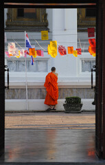 Moine bouddhiste en kesa orange, de dos, sous des drapeaux de prière, dans l'encadrure d'une porte donnant sur l'intérieur d'un temple à Bangkok. 