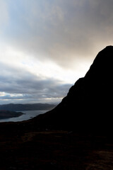 Silhouette of Bealach na Ba mountain with dramatic sunset and storm clouds with view of Loch Carron shoreline and mountain ridges beyond, Highlands of Scotland