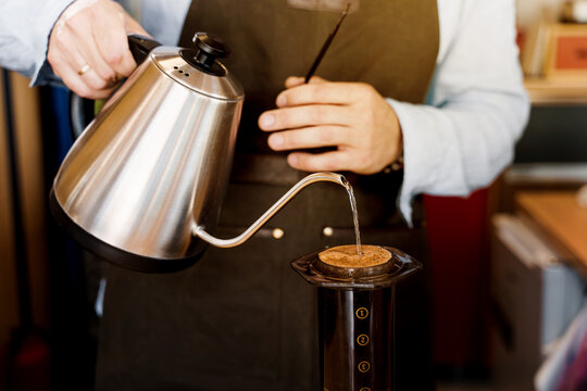 Barista holds kattle in hands and pours hot water in aeropress device with coffee. Alternative method of coffee brewing. Photo with sun light