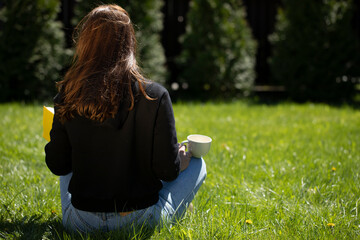Attractive brunette girl with long hair dressed in black hoodie sits on grass with book and cup of...