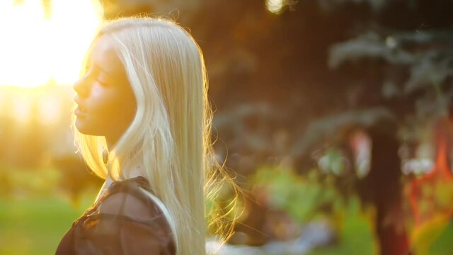Caucasian girl gracefully goes towards the setting sun past green trees in the park. Long blond fluffy hair flutters from behind, the teenager turns around, looks confidently at the camera and smiles.