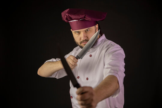 Serious chef man in uniform holds two knives crossing them as fighting. Male cook in apron standing on black background. Professional master, restaurant, food industry, kitchen cooking battle concept