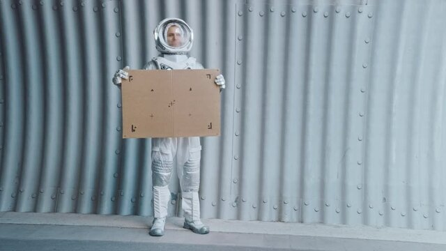 Happy Man in Spacesuit is Standing in a Metal Tube Tunnel and Holding a Carboard Mockup Sign for Your Design. Spaceman in Futuristic Suit with Technological Panel on His Hand.