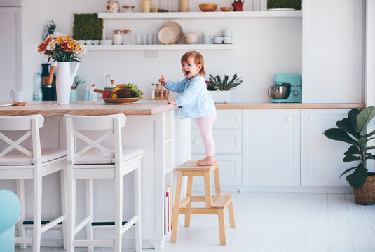 Funny Infant Baby Girl Standing On A Step Stool At The Cozy Kitchen At Home