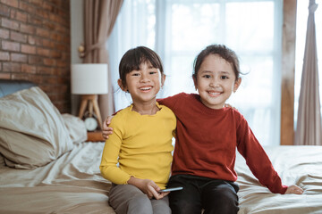 two little Asian girls laugh happily looking at the camera while sitting on the bed and holding a cellphone