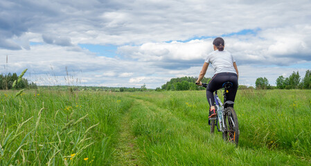 Fototapeta na wymiar Back view of Young athletic woman riding bicycle on rural road in green field or meadow in summertime against dramatic sky. Solo outdoor activities. Enjoy time alone in nature. Banner, copy space