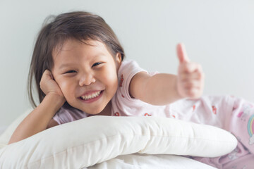 Sweet little female asian child smiling and giving a thumbs up hand gesture to the camera while laying in bed wearing pajamas posing. Closeup portrait.