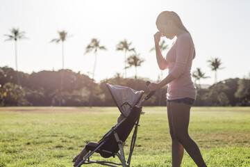 Exhausted single young mother with hand on her face pushing a stroller with her baby child at the park. Maternity stress.