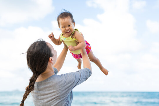 Cute baby girl smiling at the camera while her mother holds her up in the air against a blue sky on a fun beach day during summer. Family, travel and leisure.