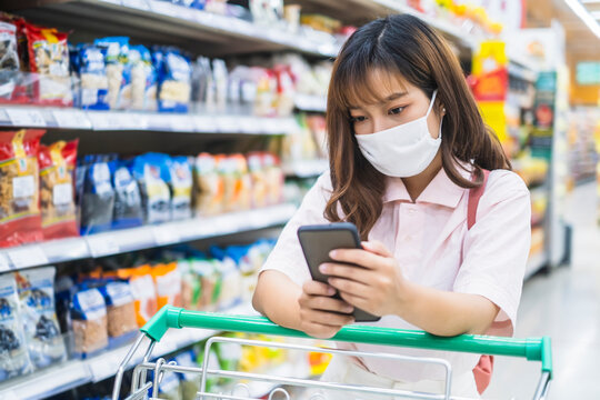 beautiful asian girl facemask protection coronavirus covid-19, smart phone checking price payment shopping buying food grocery shopping at super store market stock supply world pandemic quarantine