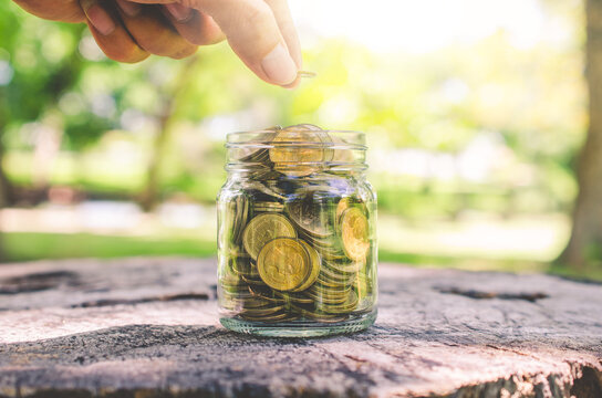 investor business man hand putting coins in jar on wood table with blur nature park background. money saving concept for financial banking and accounting.