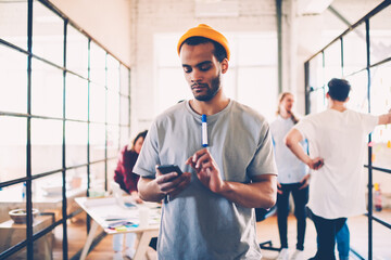 Thoughtful afro american team member reading income message on mobile during work break, dark-skinned student browsing website and checking information while colleagues talking on background