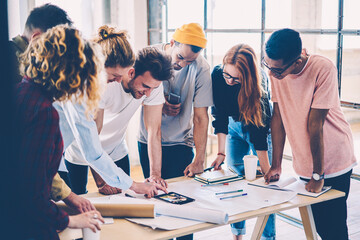 Young crew of male and female designers in casual trendy outfit communicating during conference using technology for improving planning.Skilled architects making research together drafting in studio